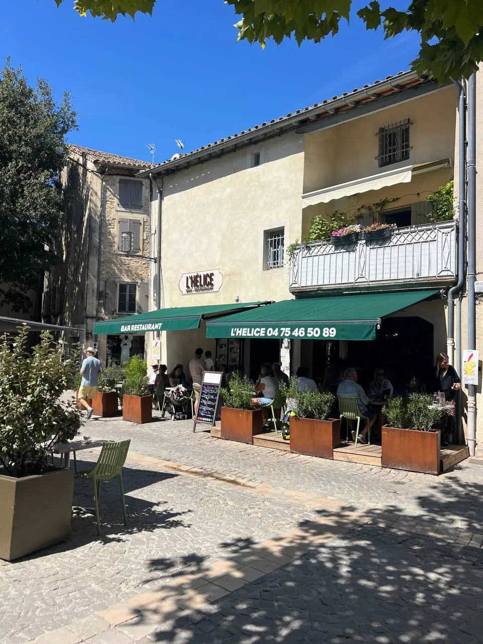 La façade de L'Hélice et sa terrasse pleine, un jour d'été à Grignan